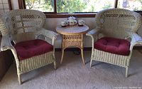 Full view of two vintage ecru painted wicker armchairs with burgundy cushions and a natural wicker round table topped with a metal teapot and two diner mugs.
