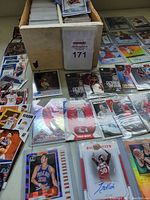 Photo showing a large box filled with basketball trading cards, visible autographed cards, jersey cards, and star player cards in protective sleeves and plastic holders spread around the box.