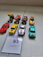 Front view of eight vintage Hot Wheels die cast cars from the 1970s arranged in two rows, showing various colors and models on a white surface with lot number 93 card visible.