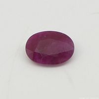 Close-up photo of oval pink-red ruby displaying faceted cut and color on a neutral background.