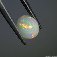 Close-up photo of the oval cabochon white opal held in clamps showing play-of-color in the stone under lighting.