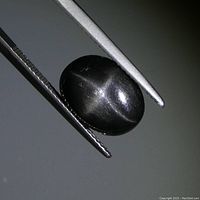 Close-up of black oval cabochon star diopside held by tweezers showing star-like reflective pattern on surface.