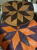 Two rounded square beanbag ottomans with leather patchwork covers, one in tan and brown leather, the other in purple and orange leather, placed stacked.