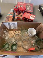 Photo showing multiple Coca-Cola glasses in a cardboard box along with collectible tins, bottles, and boxed items