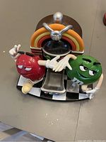 Front angle view of the M&M's candy dispenser showing both red and green M&M characters, the rainbow arch, black spherical container, and silver dispensing chute on a checkerboard base.