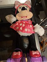 Plush Minnie Mouse electronic toy with pink polka dot dress and bow, white gloves, and black legs. Displayed on glass surface.