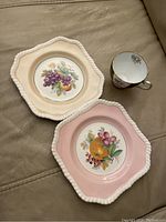 Two scalloped edge saucers with fruit designs and one floral-patterned tea cup with gold trim, placed on a tan surface.