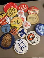 Photo showing 16 assorted vintage buttons mostly circular, with some political, promotional, and music themes.