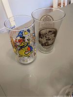 Two vintage collectible drinking glasses displayed on a table - one Disney themed with a colorful dragon wizard illustration and one Coca-Cola branded glass with sepia old-fashioned car design.