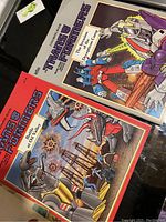 Photo shows two vintage Transformers colouring books with colorful illustrated covers depicting Transformers characters in battle scenes.