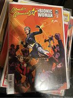 Front cover of Wonder Woman '77 Meets The Bionic Woman #2 comic book showing Bionic Woman in mid-fight stance and Wonder Woman in the background with crowd and futuristic elements.