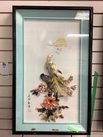 Framed 3D art with bird made of shell and ceramic perched among flowers, under glass, with dark wood frame visible.