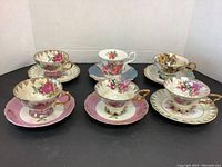 All six porcelain cups with matching saucers arranged in two rows showing floral patterns, gold trims, and opalescent finishes.