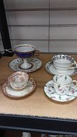 Four cups and saucers displayed on a surface, showing varied floral and gold designs including a blue-gold pedestal cup.
