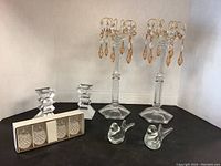 Set showing all items on black surface against white corner wall: two tall glass candle holders with hanging amber crystal prisms, two smaller geometric clear glass candle holders, two clear glass birds, boxed set of four glass pineapples.