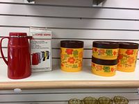 Set of four plastic canisters with colorful 70s floral and heart designs and a red coffee carafe with original box displayed on shelf.