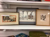 Three framed pen and ink prints of street scenes displayed on a shelf.