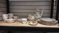 View of full set displayed on shelf with plates stacked, cups on left and gravy boat and teapot center back.