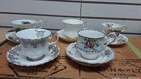 Five Royal Albert teacups each with matching saucers arranged on a wooden surface showcasing the varied floral and gold ornate patterns.