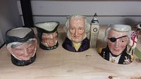 Four Toby mugs displayed on a wooden shelf, showcasing characters John Doulton, Nelson, Granny, and one unnamed.