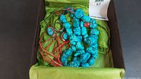 Jewelry pieces in a box with a green lining, showing large turquoise stones and red beads for the necklace, and rings with turquoise and red stones.