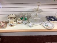 Overview of Bunnykins dishes with mugs, bowls, 3-tier cake platter, Wedgwood plate, butter dish, and drinking glasses on display shelf