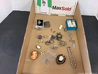 Box containing a collection of vintage jewelry including rings, brooches, and cameos spread inside a cardboard box.