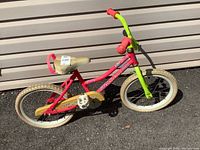 Side view of the Fisher Price children’s bicycle showing the pink and green frame, white tires that are flat, beige saddle seat, and yellow-orange chain guard.