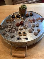 Top down view of twelve pairs of costume earrings and three pins arranged on a round metal tray, showing variety of styles and materials.