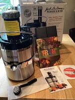 Front view of stainless steel and black Jack LaLanne Power Juicer Ultimate with juice spout, recipe booklet, instruction manual and DVD in front, and original box in background