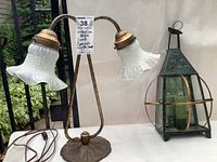 Full view of the Victorian style metal lamp with two frosted glass flower-shaped shades, showing the decorative legs and wired cord, placed on a white table with a background of plants and wall.