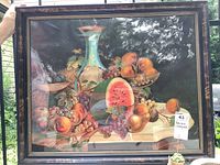 Full view of the framed antique lithograph depicting a still life of summer fruits and a glass decanter, with visible reflections on glass.