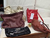Five purses and bags arranged on table including black pleated clutch, maroon Fossil leather bag, gold metallic The Limited bag, brown Soprano leather bag, and red BE organizer bag