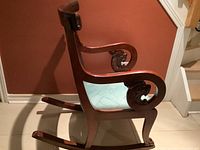 Side profile photo showing the wood frame of the rocking chair with detailed lion head carving on the armrest and side panel, dark mahogany finish, and curved rockers.