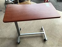 Overbed table with wood grain surface in rectangular shape, metal adjustable base, and four caster wheels shown flat.