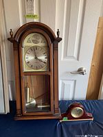 Photo showing both clocks side by side: grandfather style clock on left, tabletop clock on right on blue surface, near door