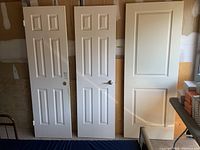 Three white interior doors leaning against a wall in a garage or storage area. Two six-panel hollow core doors and one two-panel sliding door panel are shown.