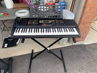 Full front view of Yamaha PSR-225 electronic keyboard on X-style stand outside garage, with power adapter on left side of keyboard.