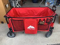 Front view of red Ozark Trail folding wagon on a concrete floor, showing mesh storage pockets with central flap and handle on side.