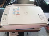 Top-left angle view of HP DeskJet all-in-one printer showing general condition, stickers on top, and basic design.