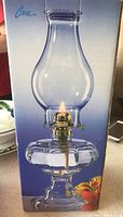 Front view of the Crisa oil lamp in its original box depicting the glass and metal oil lamp with a flame lit.