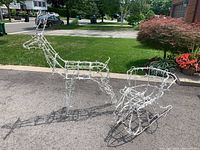Full side view of white metal wire frame reindeer and sleigh set outdoors on pavement with green grass and sidewalk visible in background.