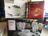 Photo showing boxed porcelain dish set with mugs, plates, and bowls from Canvas brand, Stokes porcelain salad bowl set in box, and silver-plated wire basket in box.