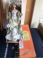 Porcelain doll seated inside original box, showing full outfit and facial features along with certificate of authenticity and box branding.