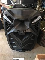 Front view of the black Sylvania SPA124 Bluetooth tailgate speaker showing angular front design with mesh grille and two white speaker covers. Wired handheld microphone on top.