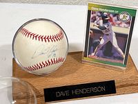 Close-up of the autographed baseball in clear plastic case next to Dave Henderson baseball card on wooden stand with name plaque.