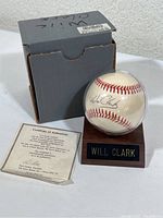 Baseball autographed by Will Clark displayed in clear protective case on wooden stand with nameplate, accompanied by COA and original box.