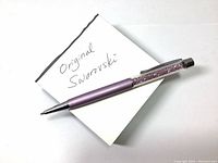 Genuine Swarovski ballpoint pen placed on a white surface with a handwritten note stating 'Original Swarovski'. Pen upper section filled with purple violet mini crystal pieces inside clear acrylic shell.