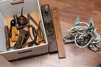 Box containing various vintage hand tools including oil can, hand drill, pliers, and measuring devices alongside wooden level and hand plane on floor.