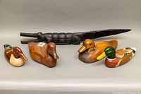 Photo showing five handcrafted wooden sculptures: a dark elongated crocodile with segmented body and four bird decoys in various browns and greens.